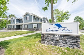  A sign in front of the facility reading "Sunrise Detox - A New Beginning," placed on a stone base with neatly landscaped greenery.