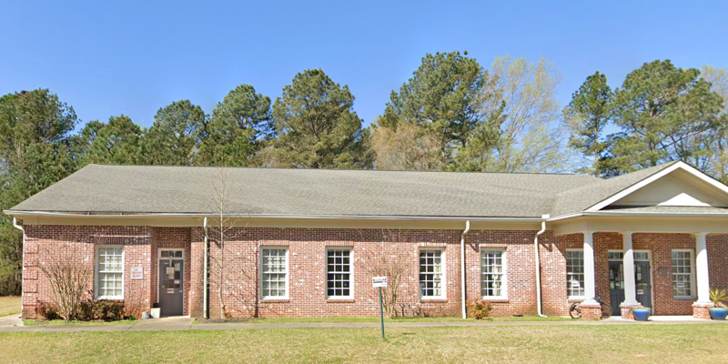 A single-story red brick building with white columns at the entrance, surrounded by green trees and a well-maintained lawn, serving as the Advantage Behavioral Health Systems Walton County Mental Health Center.