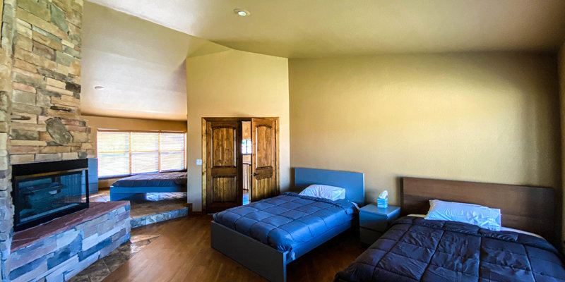 Comfortable bedroom with two beds, a fireplace, wood flooring, natural light, and stone and wood accents