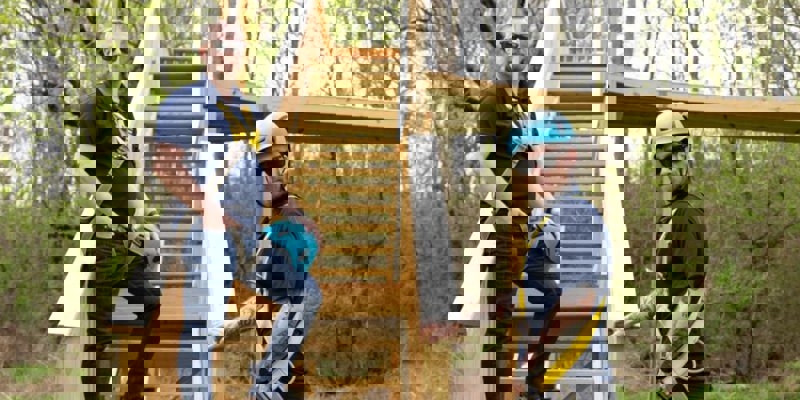 Two staff members in safety harnesses standing by the wooden stairs of a ropes course in a forested area