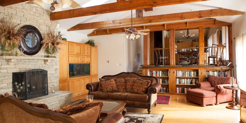 Warm-toned living room with wooden beams, fireplace, plush seating, and bookshelves
