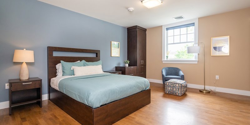  A contemporary and neatly arranged bedroom with a plush bed, wooden flooring, a bedside lamp, and a cozy armchair, creating a relaxing ambiance.